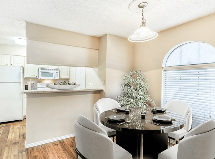 Virtually staged dining room with round table and chairs, wood style flooring, hanging light fixture, large window with blinds, and kitchen with breakfast bar in background